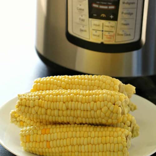 Four ears of corn on a white plate in front of an Instant Pot pressure cooker.
