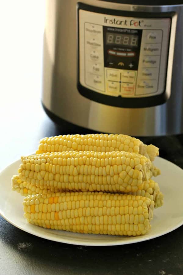 Four ears of corn on a white plate in front of an Instant Pot pressure cooker.