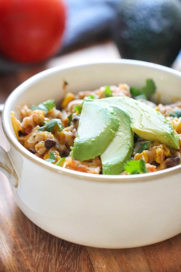 A white bowl filled with a rice and bean mixture, topped with slices of avocado and garnished with chopped herbs. A tomato and avocado are blurred in the background.