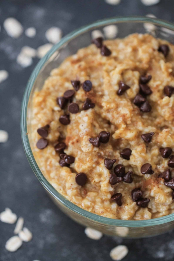 A glass bowl filled with oatmeal topped with chocolate chips. Scattered oats and a blurred can in the background.