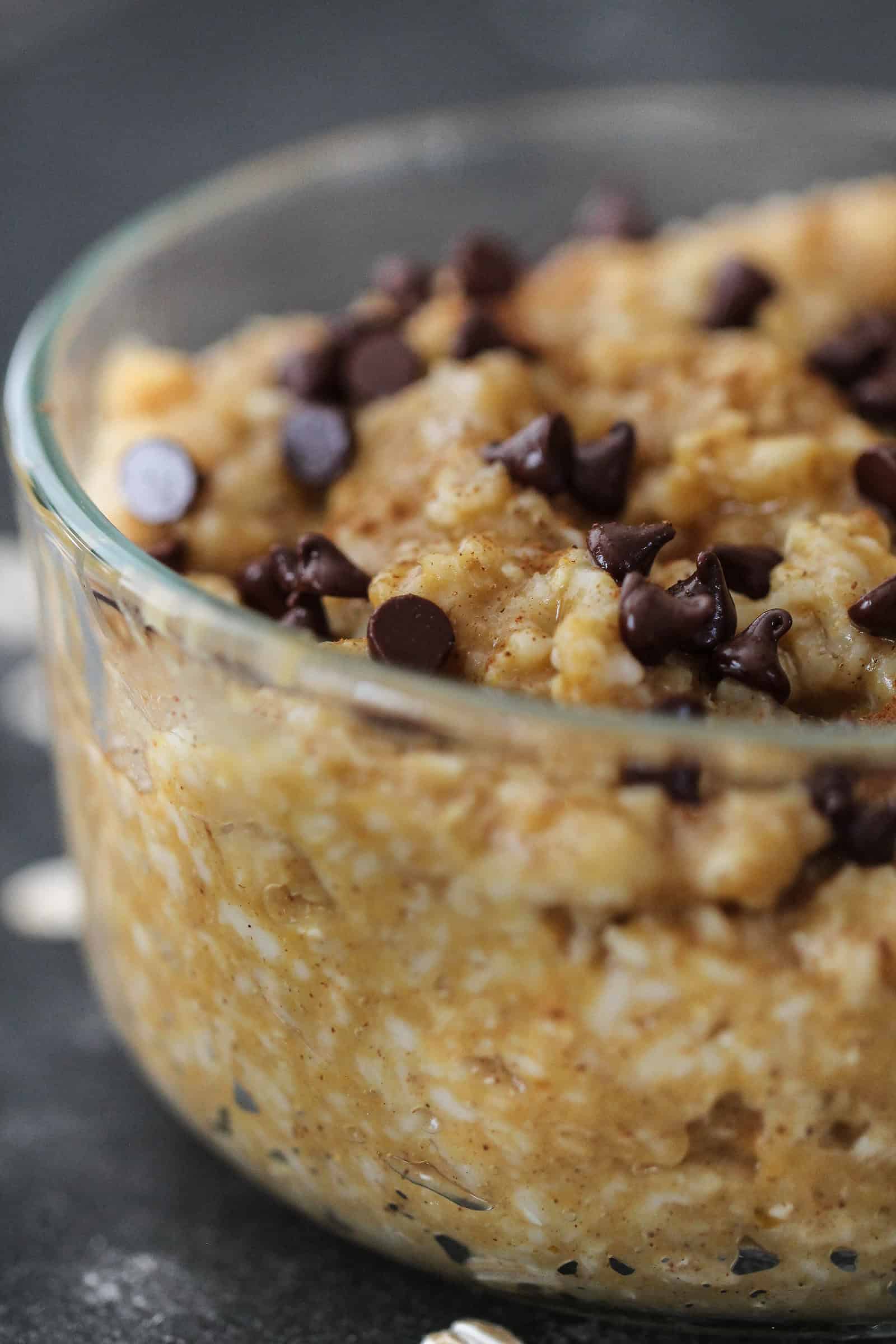 A glass bowl filled with oatmeal topped with chocolate chips. Scattered oats and a blurred can in the background.