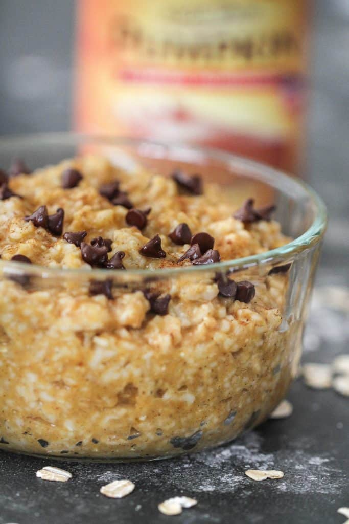 A glass bowl filled with oatmeal topped with chocolate chips. Scattered oats and a blurred can in the background.