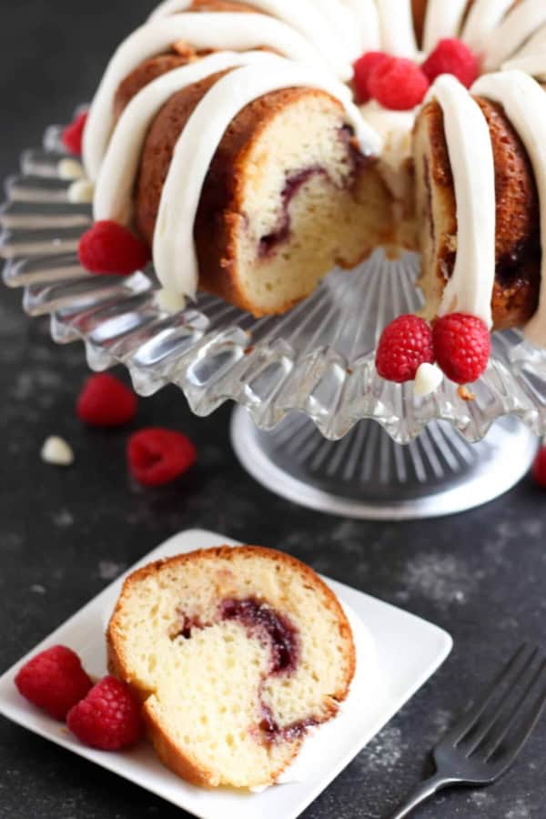 White Chocolate Raspberry Bundt Cake on a cake stand with one slice below it on a plate with fresh raspberries.