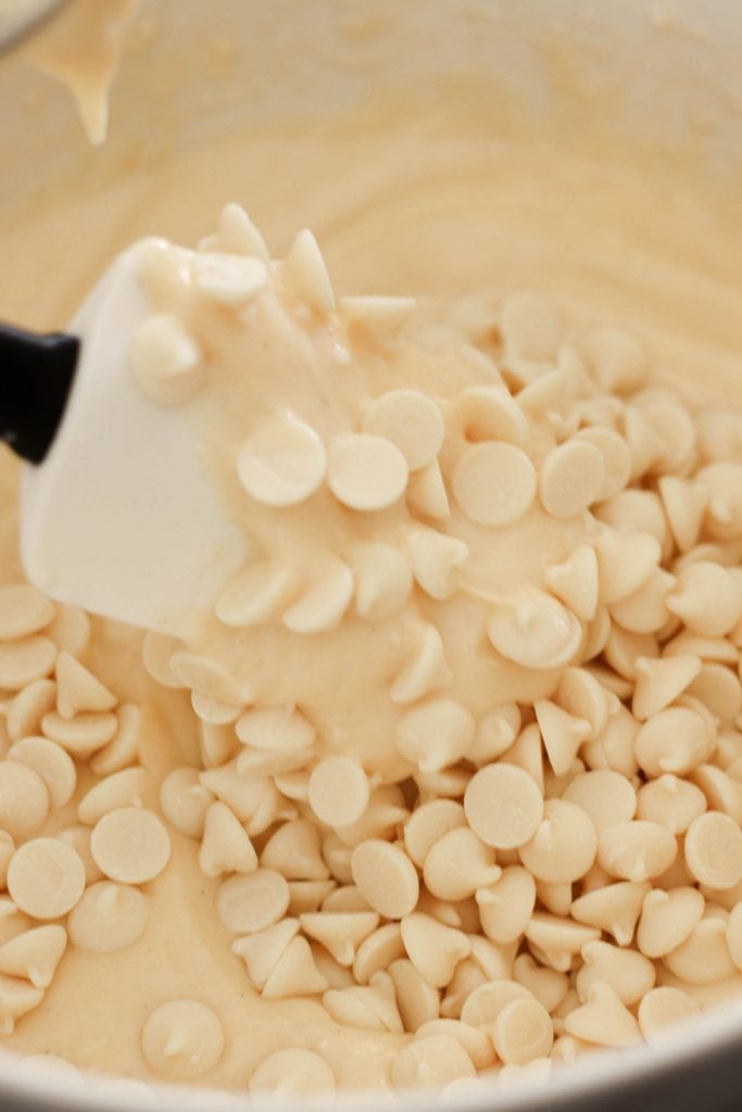 A rubber spatula stirring white chocolate chips into a mixing bowl of cake batter.