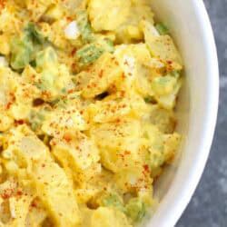 A close-up of a bowl of potato salad with chopped potatoes, green onions, and a creamy dressing, sprinkled with paprika.