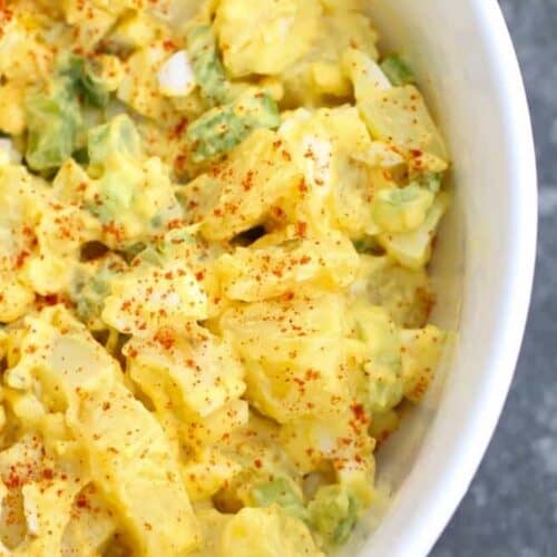 A close-up of a bowl of potato salad with chopped potatoes, green onions, and a creamy dressing, sprinkled with paprika.