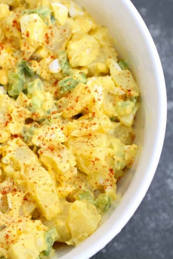 A close-up of a bowl of potato salad with chopped potatoes, green onions, and a creamy dressing, sprinkled with paprika.