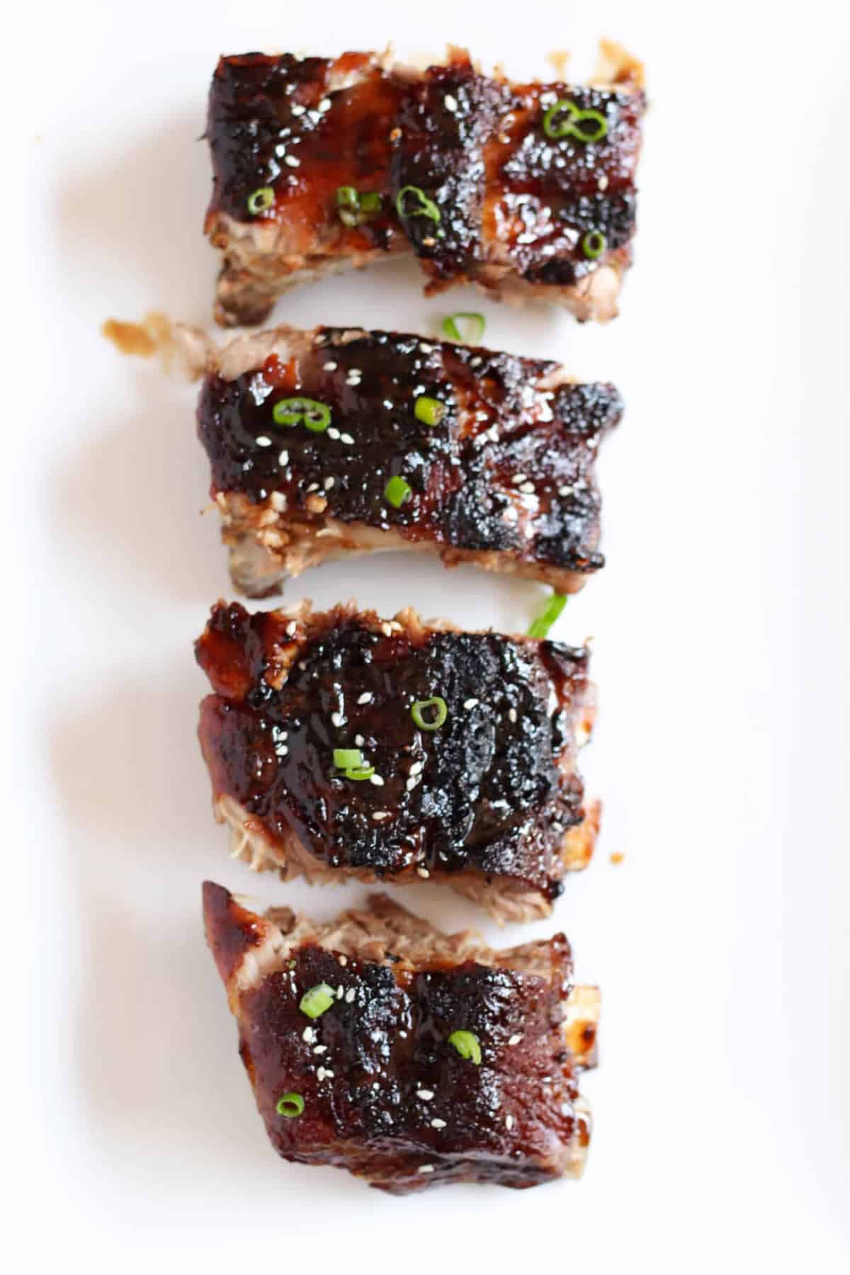 Four pieces of glazed, cooked ribs are arranged in a vertical line on a white surface, garnished with small green herbs.