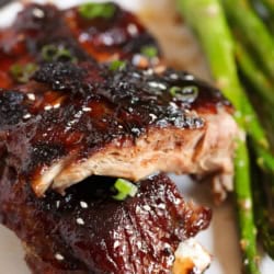 Close-up of glazed barbecued ribs garnished with green onions and sesame seeds, served with roasted asparagus on a white plate.