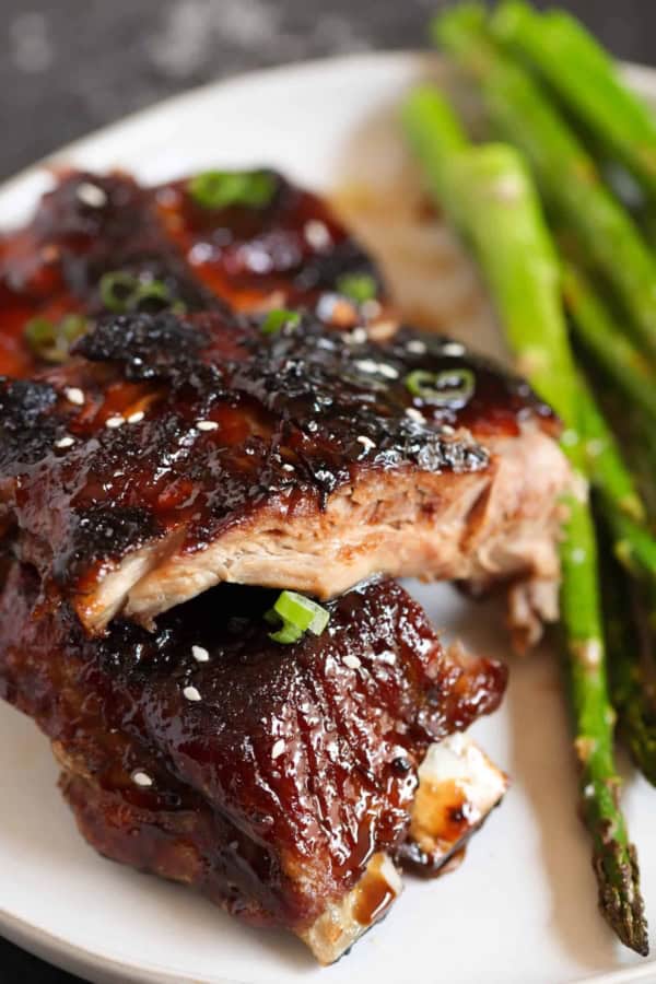 Close-up of glazed barbecued ribs garnished with green onions and sesame seeds, served with roasted asparagus on a white plate.