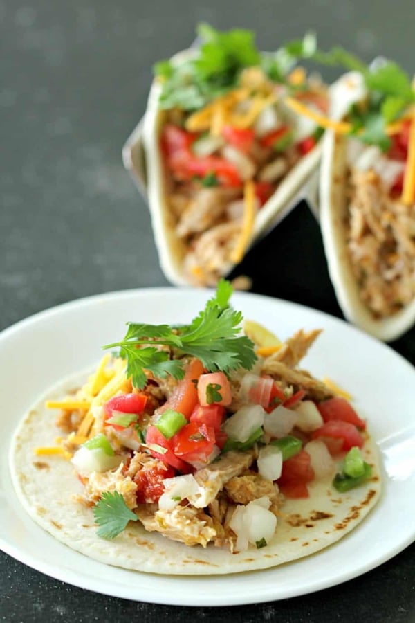 A taco shell filled with pulled chicken, diced tomatoes, onions, chopped green peppers, cilantro, and shredded cheese on a white plate. Another taco can be seen in the background.