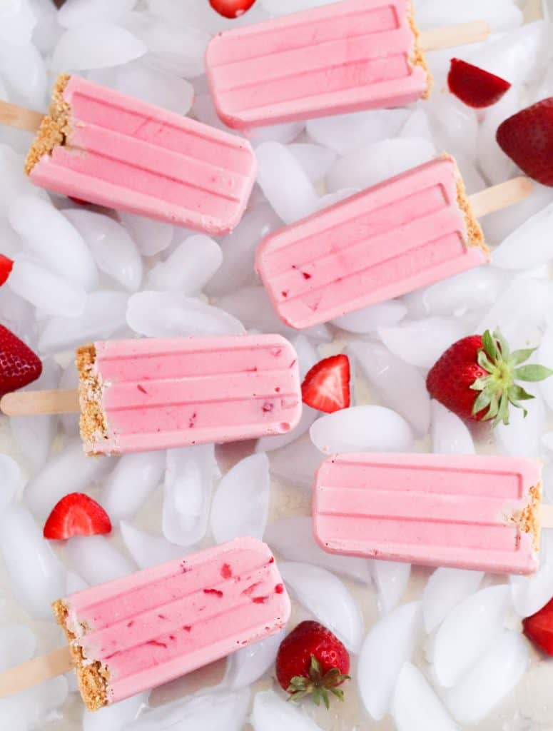 Strawberry Cheesecake Popsicles laying on ice cubes with fresh strawberries.