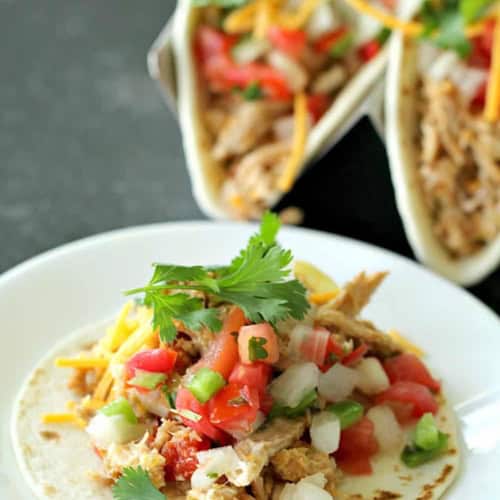 A taco shell filled with pulled pork, diced tomatoes, onions, chopped green peppers, cilantro, and shredded cheese on a white plate. Another taco can be seen in the background.
