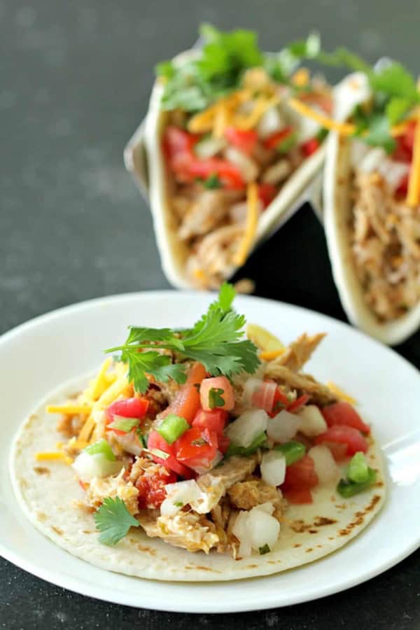 A taco shell filled with pulled pork, diced tomatoes, onions, chopped green peppers, cilantro, and shredded cheese on a white plate. Another taco can be seen in the background.