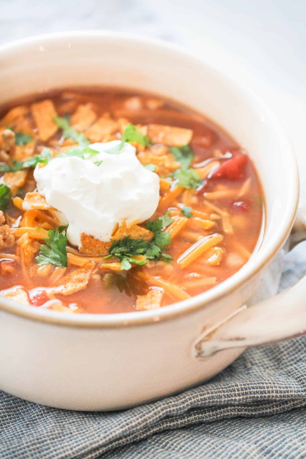 A bowl of tomato-based soup topped with a dollop of sour cream, grated cheese, cilantro, tortilla strips, and garnished with chopped cilantro.
