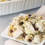 A square white plate with a serving of chicken and brown rice topped with black beans and corn, with a white dish containing more of the same food in the background.