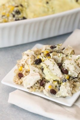 A square white plate with a serving of chicken and brown rice topped with black beans and corn, with a white dish containing more of the same food in the background.