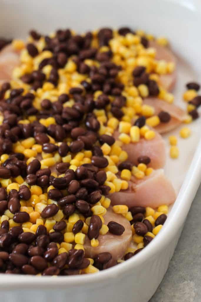 Raw chicken pieces in a white baking dish topped with black beans and corn kernels, ready to be cooked.