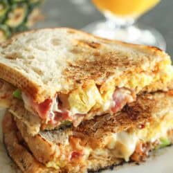 Grilled sandwich with melted cheese, egg, and bacon on a plate, accompanied by a blurred glass of orange juice in the background.