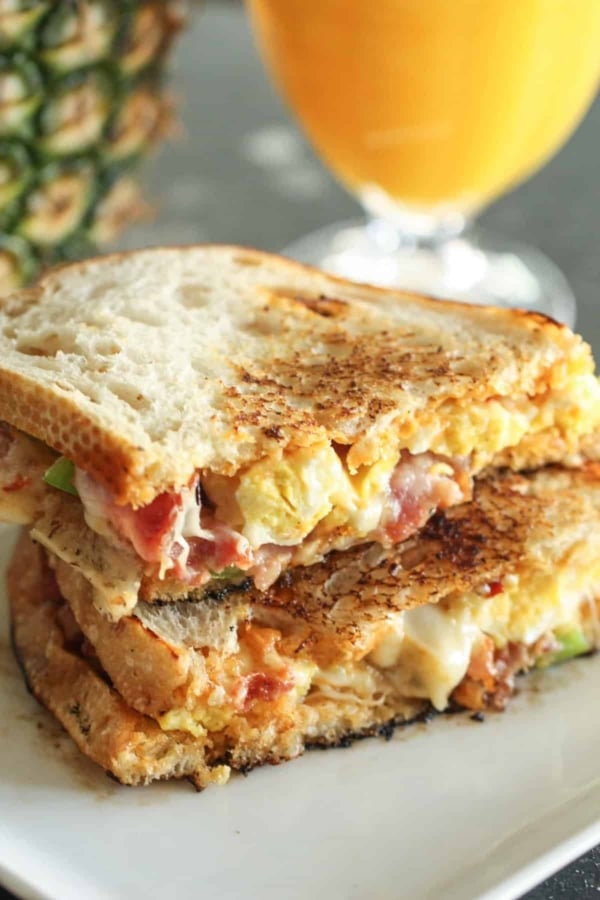 Grilled sandwich with melted cheese, egg, and bacon on a plate, accompanied by a blurred glass of orange juice in the background.