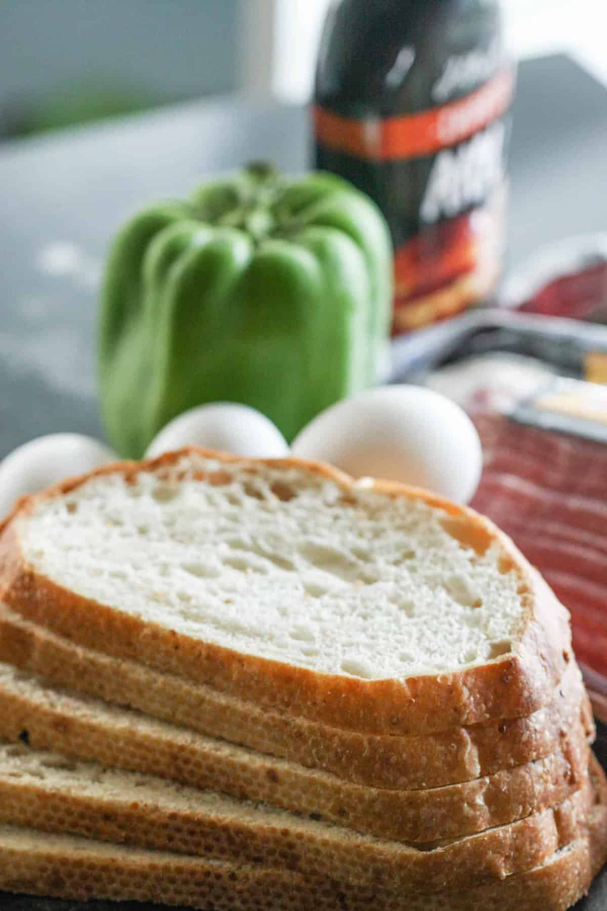 Slices of bread, a green bell pepper, and three eggs are on a table with a jar and cloth in the background.