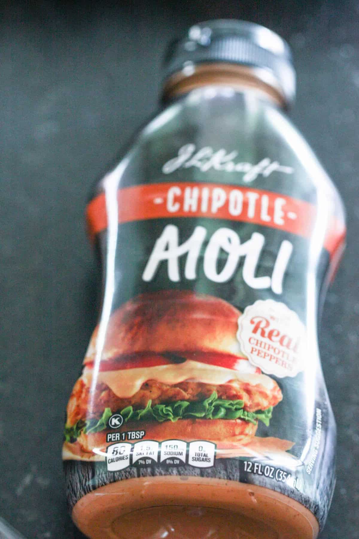 Bottle of chipotle aioli sauce on a dark surface. The label shows a burger image and lists nutritional information, product weight as 12 fl oz (355 ml), and mentions real chipotle peppers.