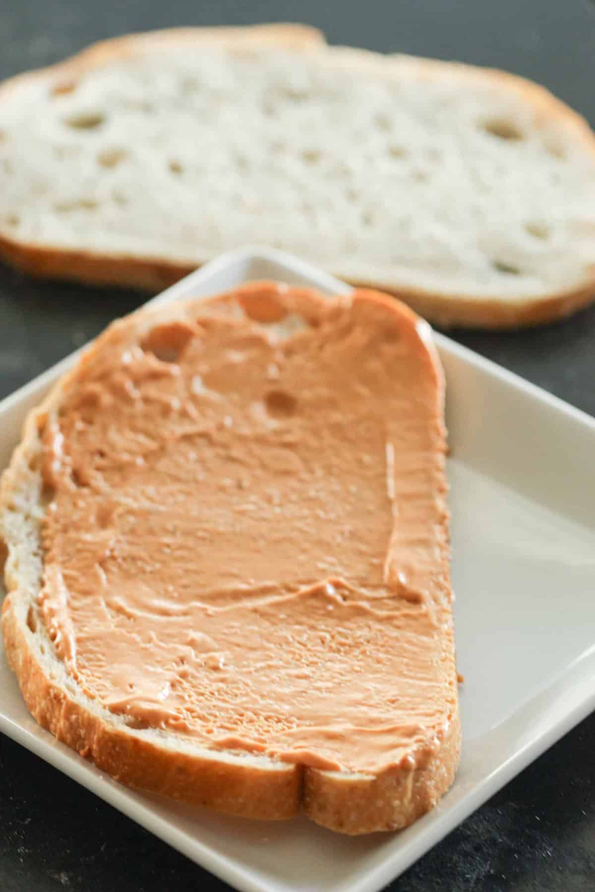 Slice of bread with peanut butter on a white plate, with another plain slice in the background.