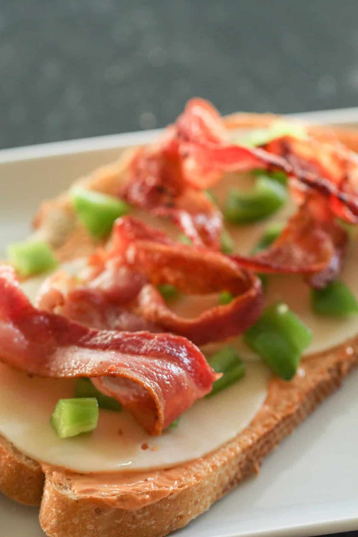 Open-faced sandwich with crispy bacon, sliced cheese, and diced green bell peppers on toast, served on a white plate.
