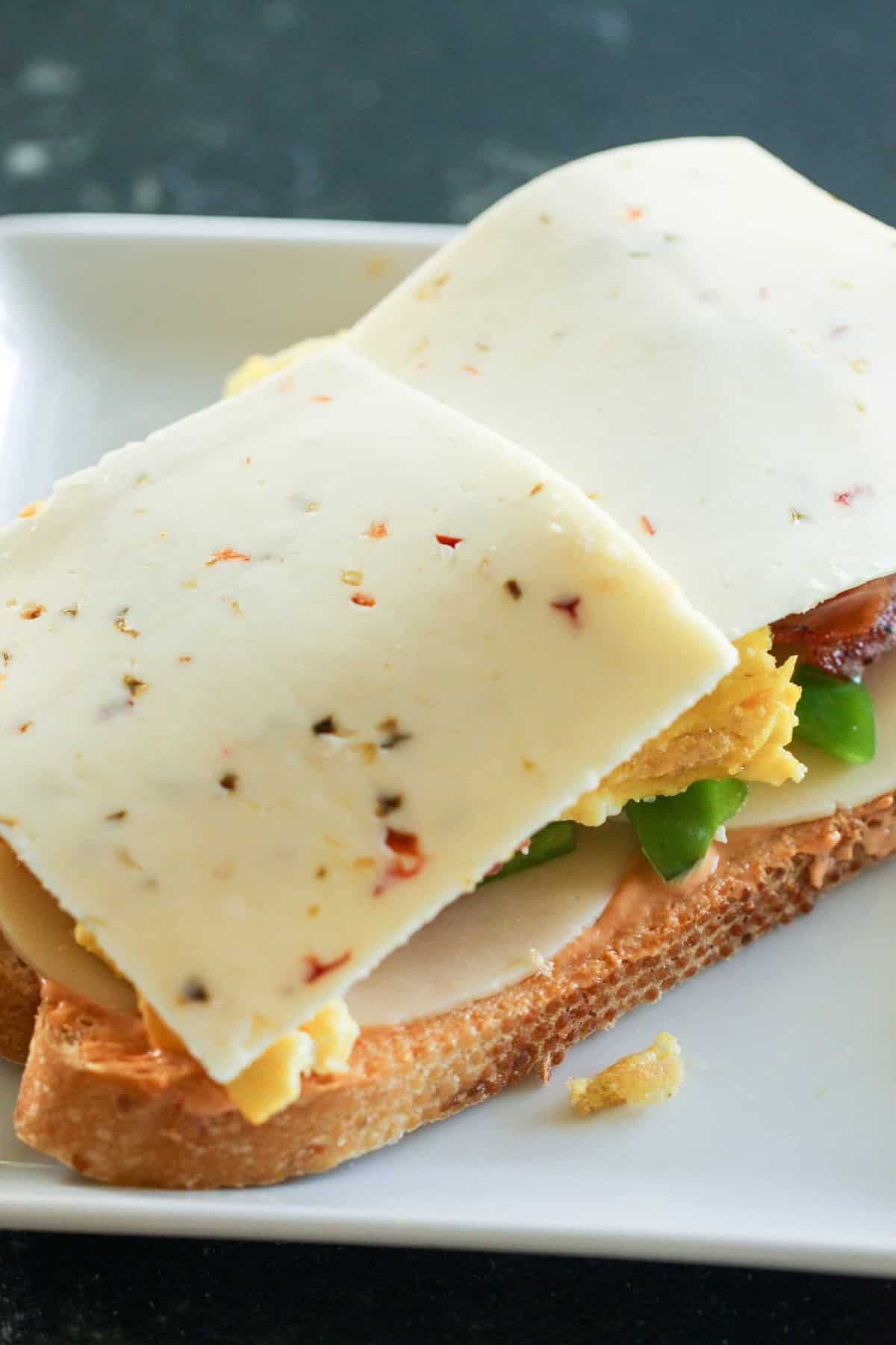A sandwich with sliced cheese containing pepper flakes, layers of scrambled eggs, green bell peppers, and turkey on multigrain bread, placed on a white plate.