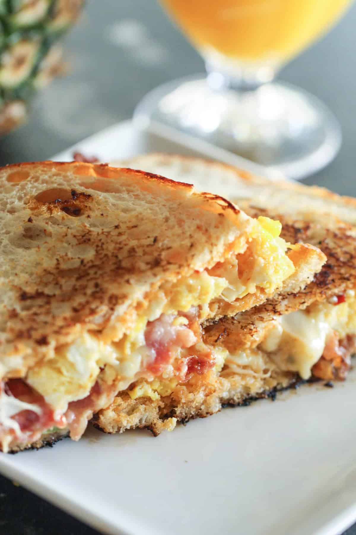 Grilled sandwich with egg, bacon, and cheese, sliced in half on a plate, with a glass of orange juice in the background.