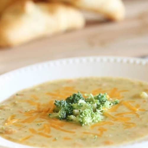 A bowl of broccoli cheddar soup garnished with shredded cheese and broccoli florets, with bread in the background.
