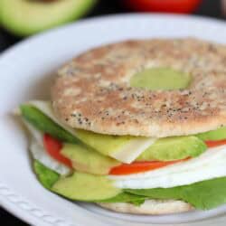 A sandwich on a seeded thin bun with lettuce, sliced tomato, avocado, cheese, and a fried egg. Background shows half an avocado and a tomato.
