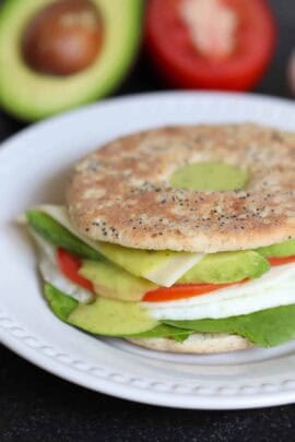 A sandwich on a seeded thin bun with lettuce, sliced tomato, avocado, cheese, and a fried egg. Background shows half an avocado and a tomato.