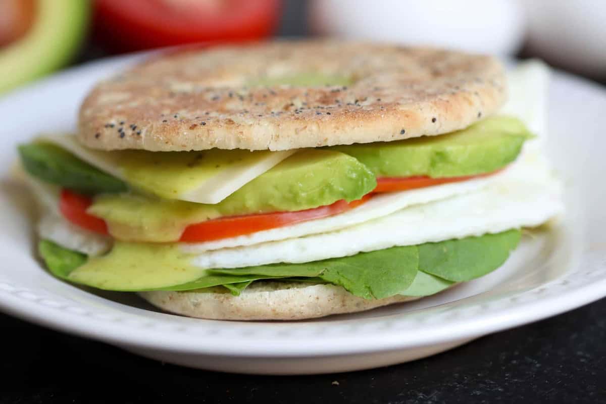A sandwich with spinach, tomato, egg white, avocado, cheese, and sauce in a thin, round bread on a white plate.