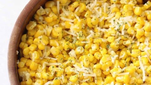 A wooden bowl filled with cooked corn kernels, topped with shredded cheese and sprinkled with herbs.