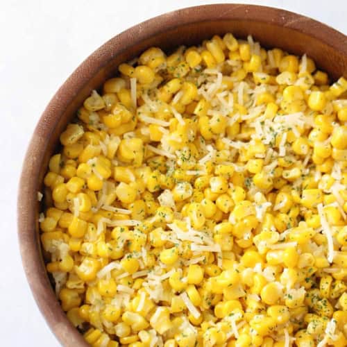 A wooden bowl filled with cooked corn kernels, topped with shredded cheese and sprinkled with herbs.