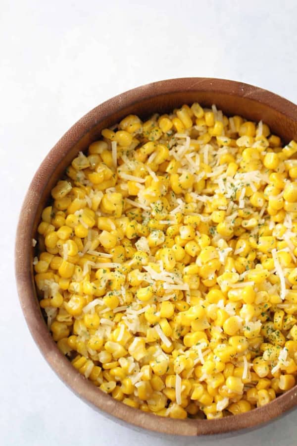 A wooden bowl filled with cooked corn kernels, topped with shredded cheese and sprinkled with herbs.