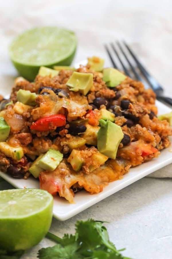 A square white plate with a serving of quinoa casserole topped with avocado chunks, surrounded by lime halves and a fork in the background.
