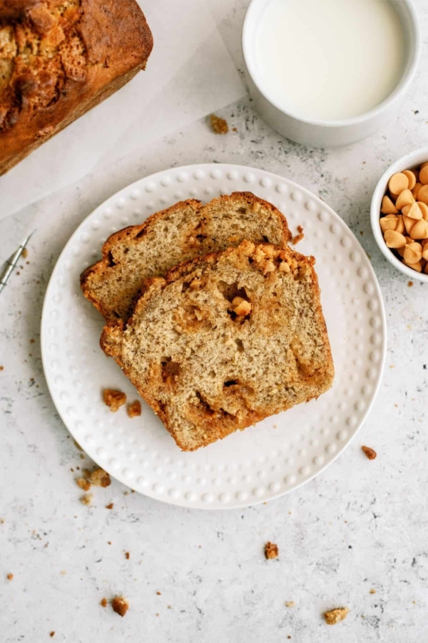 Two slices of Banana Bread on a small white plate with a cup of milk and a small cup of butterscotch chips on the side.