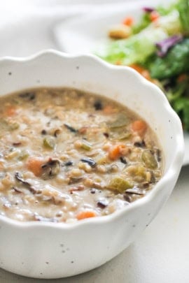 A bowl of creamy soup with visible chunks of vegetables and grains, accompanied by a side salad in the background.