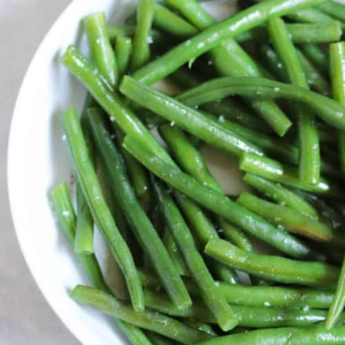 A white bowl filled with cooked green beans, lightly seasoned with salt.