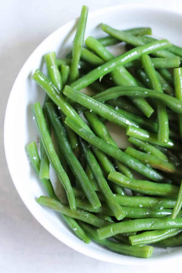 A white bowl filled with cooked green beans, lightly seasoned with salt.