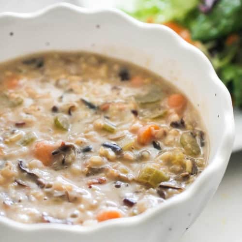 A white bowl filled with creamy wild rice and vegetable soup, with a plate of mixed green salad blurred in the background.
