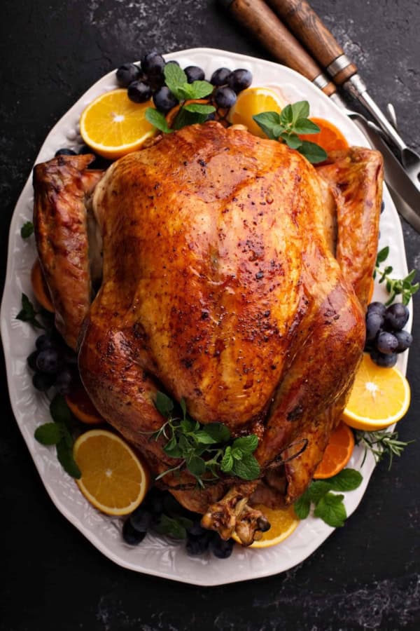 Roast turkey on a white platter, garnished with orange slices, grapes, and fresh herbs, with carving utensils nearby on a dark surface.