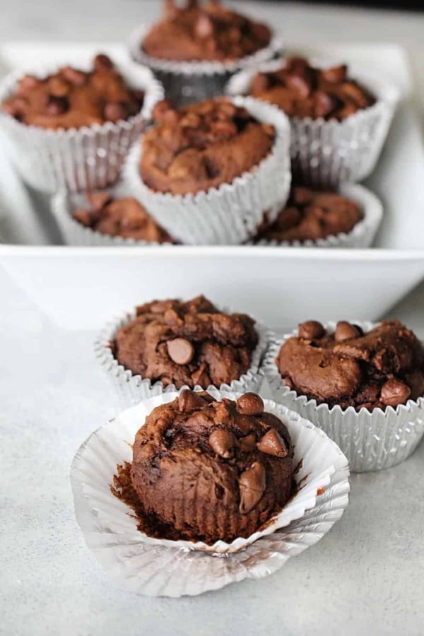 A white tray with multiple chocolate muffins in silver foil liners, with three muffins in the foreground and several more stacked in the background.
