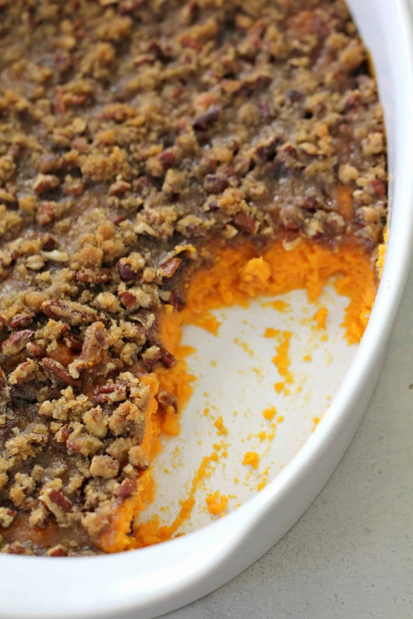 A baking dish with a pecan-topped sweet potato casserole, partially eaten, revealing orange sweet potato filling beneath the crumbly brown topping.