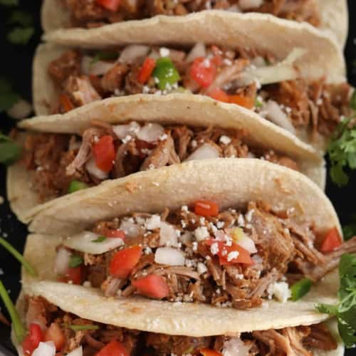 Five shredded meat tacos in soft corn tortillas, topped with diced tomatoes, onions, cilantro, and crumbled cheese, arranged closely on a dark surface.