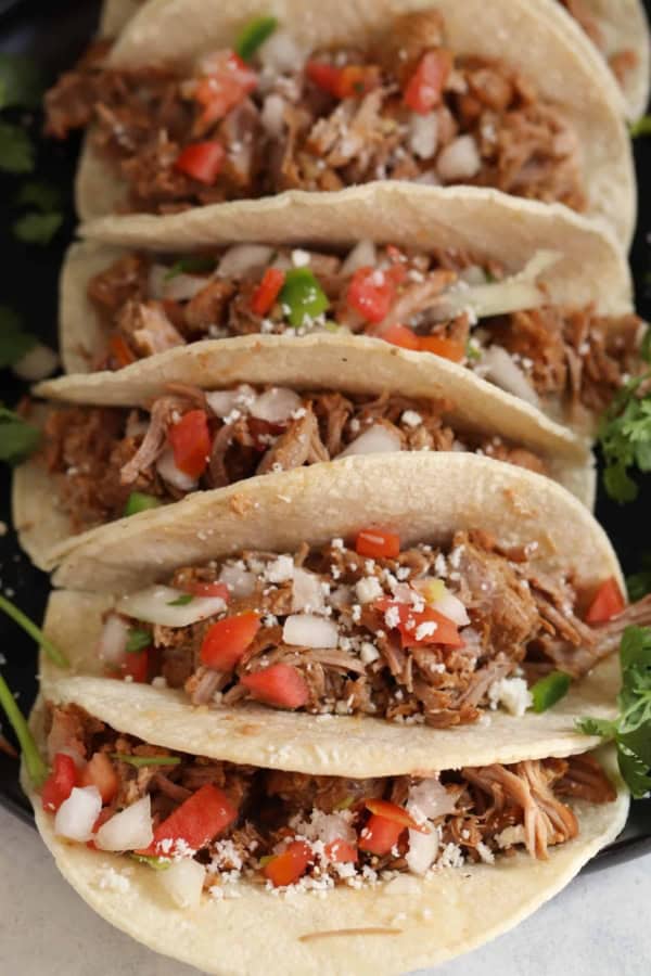 Five shredded meat tacos in soft corn tortillas, topped with diced tomatoes, onions, cilantro, and crumbled cheese, arranged closely on a dark surface.