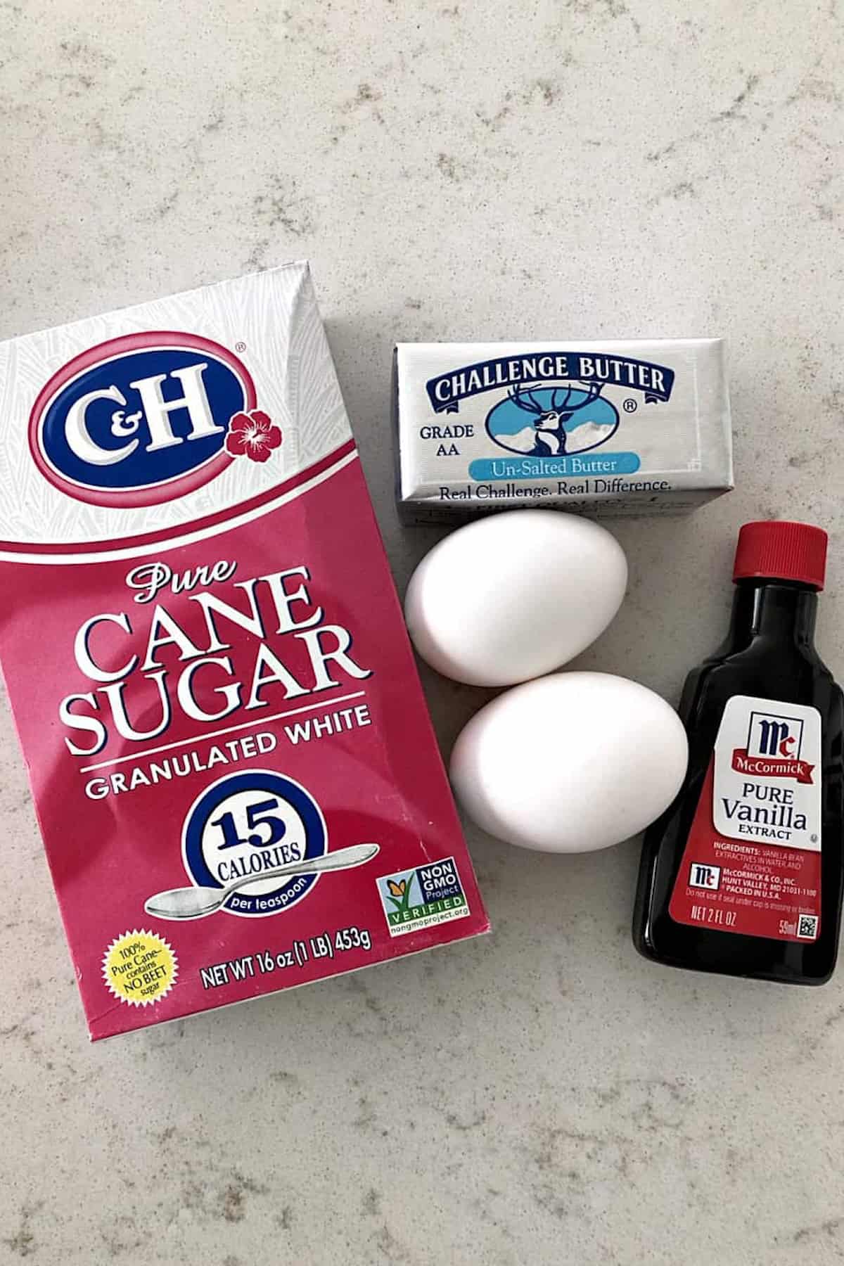 A box of granulated cane sugar, a stick of unsalted butter, two white eggs, and a bottle of pure vanilla extract on a light countertop.