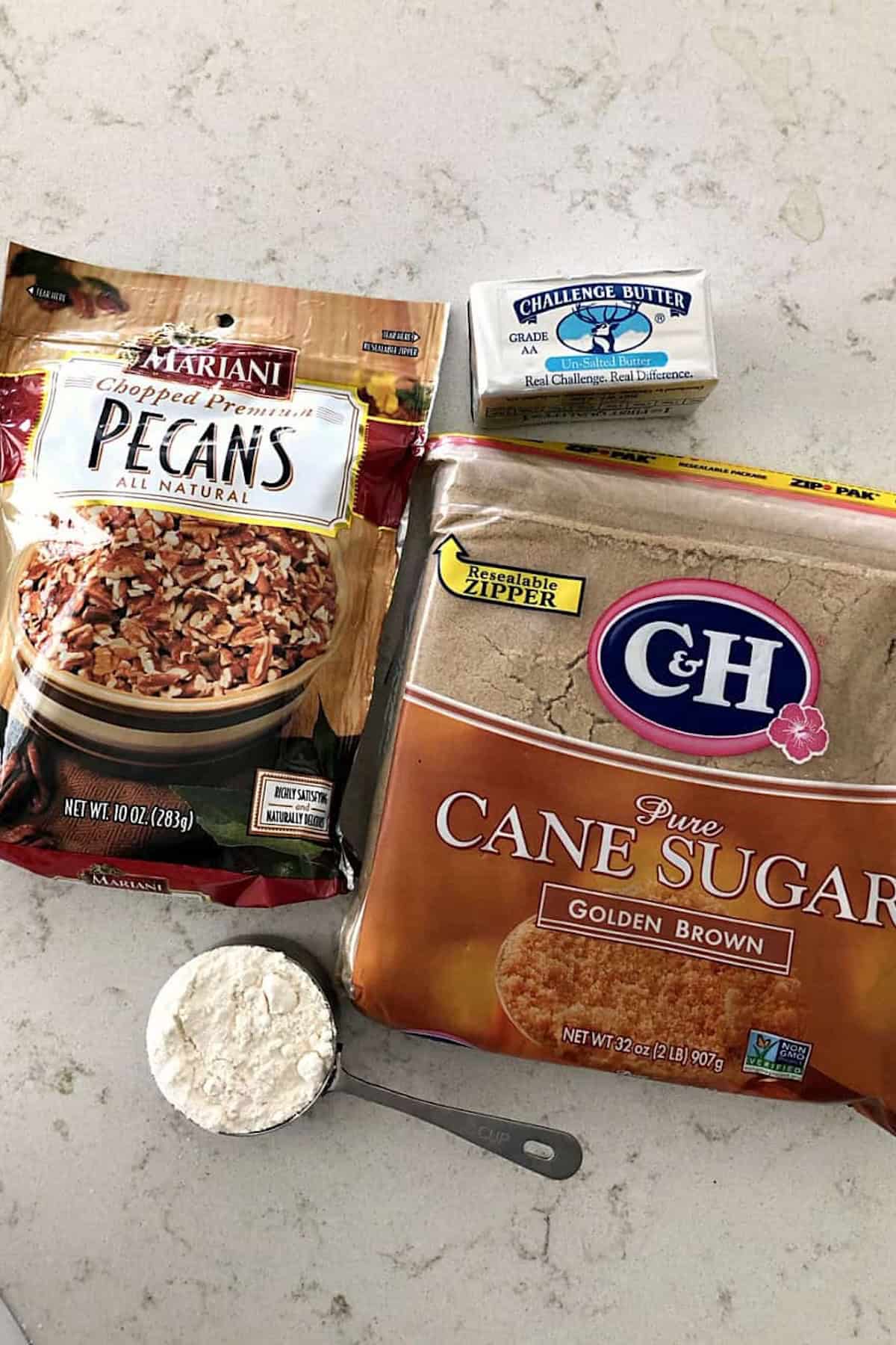 A package of chopped pecans, a box of butter, a bag of brown sugar, and a measuring cup with flour on a white countertop.